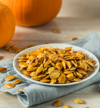 Golden roasted pumpkin seeds piled on a white plate over a blue-gray linen napkin, with fresh pumpkins in the background.